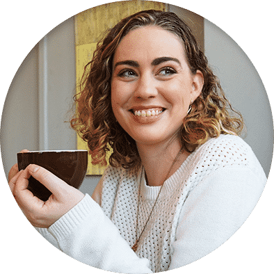 Color image of Jill Knobeloch, a woman with shoulder length curly blond hair and wearing a white sweater and gold necklace, holding a cup of coffee and smiling and looking away from the camera.